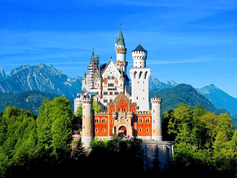 Das Schloss Neuschwanstein mit seinen Türmen liegt im Sonnenschein. Im Hintergrund sind Berge, Wald und blauer Himmel zu sehen.
