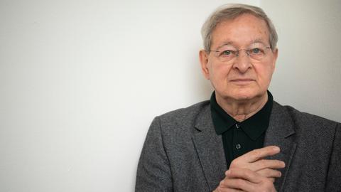 Der ungarische Schriftsteller Péter Nádas im Porträt. Er hält seine Hände verschränkt, hat graue Haaare und eine Brille. Er trägt ein graues Jackett zum schwarzen Hemd. 