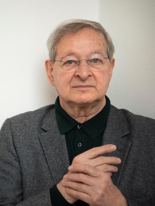 Der ungarische Schriftsteller Péter Nádas im Porträt. Er hält seine Hände verschränkt, hat graue Haaare und eine Brille. Er trägt ein graues Jackett zum schwarzen Hemd. 