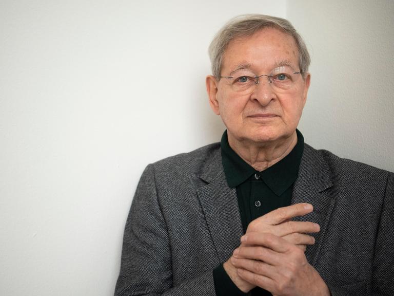 Der ungarische Schriftsteller Péter Nádas im Porträt. Er hält seine Hände verschränkt, hat graue Haaare und eine Brille. Er trägt ein graues Jackett zum schwarzen Hemd. 