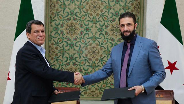 Ahmed al-Scharaa und Maslum Abdi geben sich die Hand, im Hintergrund sind Flaggen Syriens zu sehen. 