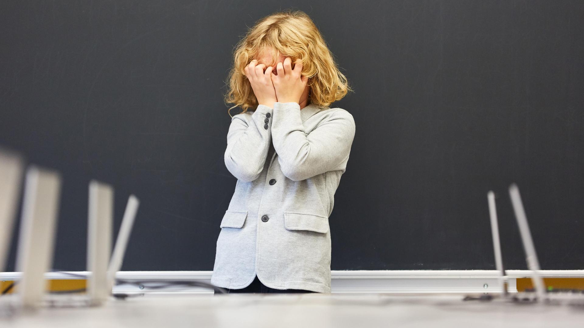 Ein Schüler steht vor einer Tafel und hält sich die Augen mit seinen Händen zu.