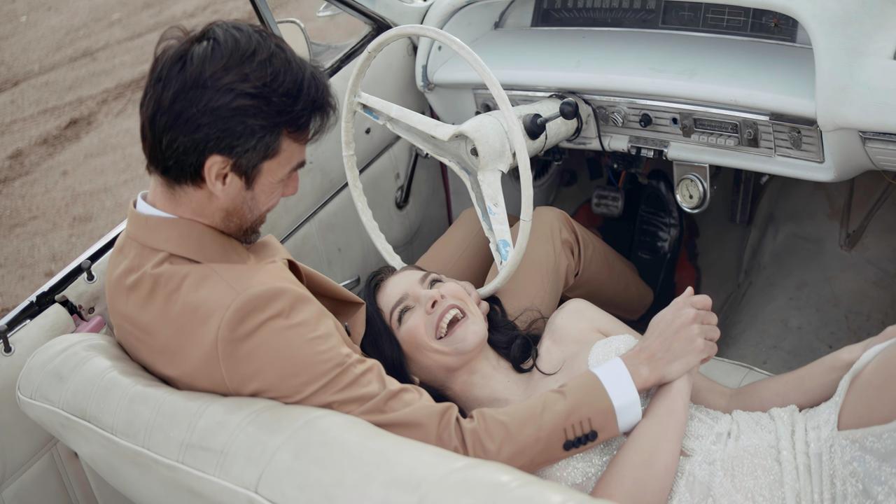 Glückliche lächelnde Frau, die in einem weißen Kleid, vermutlich einem Hochzeitskleid, auf den Knien ihres vermutlich frisch vermählten Mannes im Cabrio liegt. Er sitzt am Steuer und hält liebevoll ihre Hand.