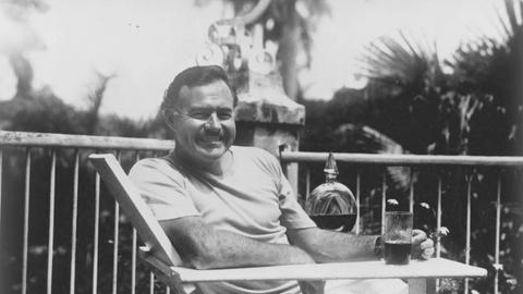 Hemingway sitzt mit einem Drink auf der Terrasse und lächelt in die Kamera.