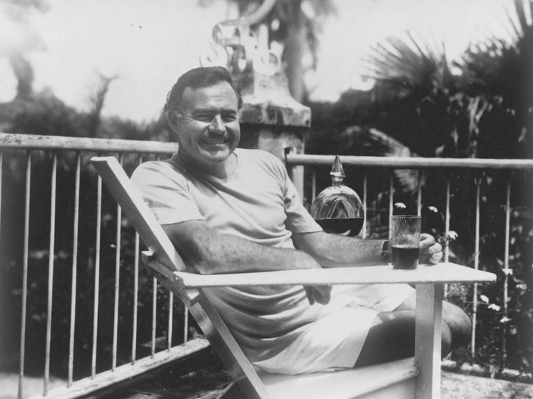 Hemingway sitzt mit einem Drink auf der Terrasse und lächelt in die Kamera.