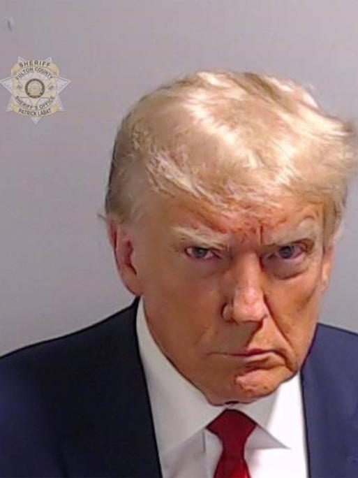 Donald Trump schaut grimmig in die Kamera. In der linken oberen Ecke ein Logo der Polizei. 