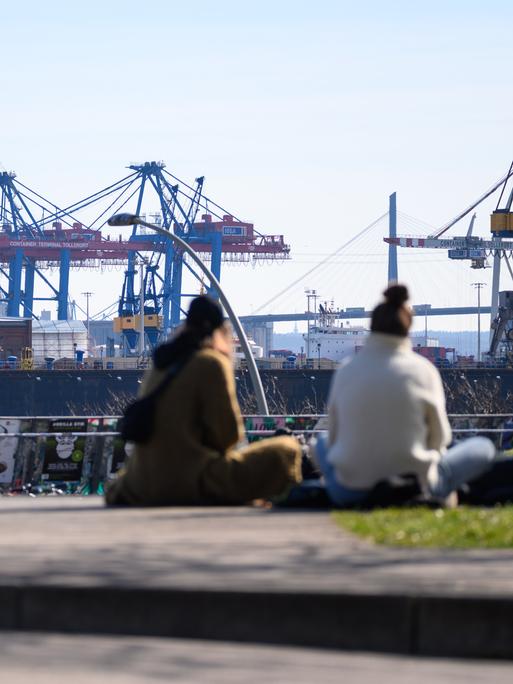 Passanten sitzen im Sonnenschein im Park Fiction auf St. Pauli gegenüber zahlreichen Hafenkränen und Docks.