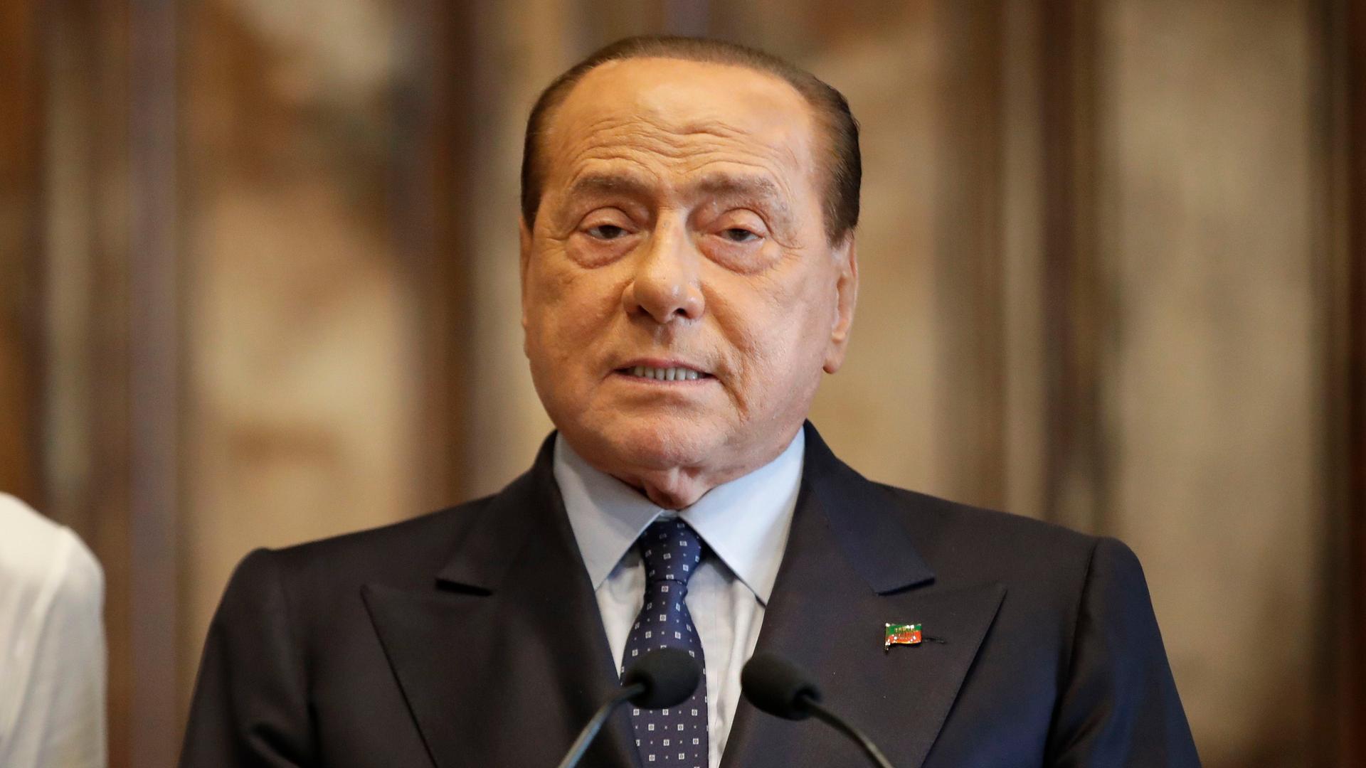 Silvio Berlusconi steht vor Mikrofonen. Er trägt einen Anzug.