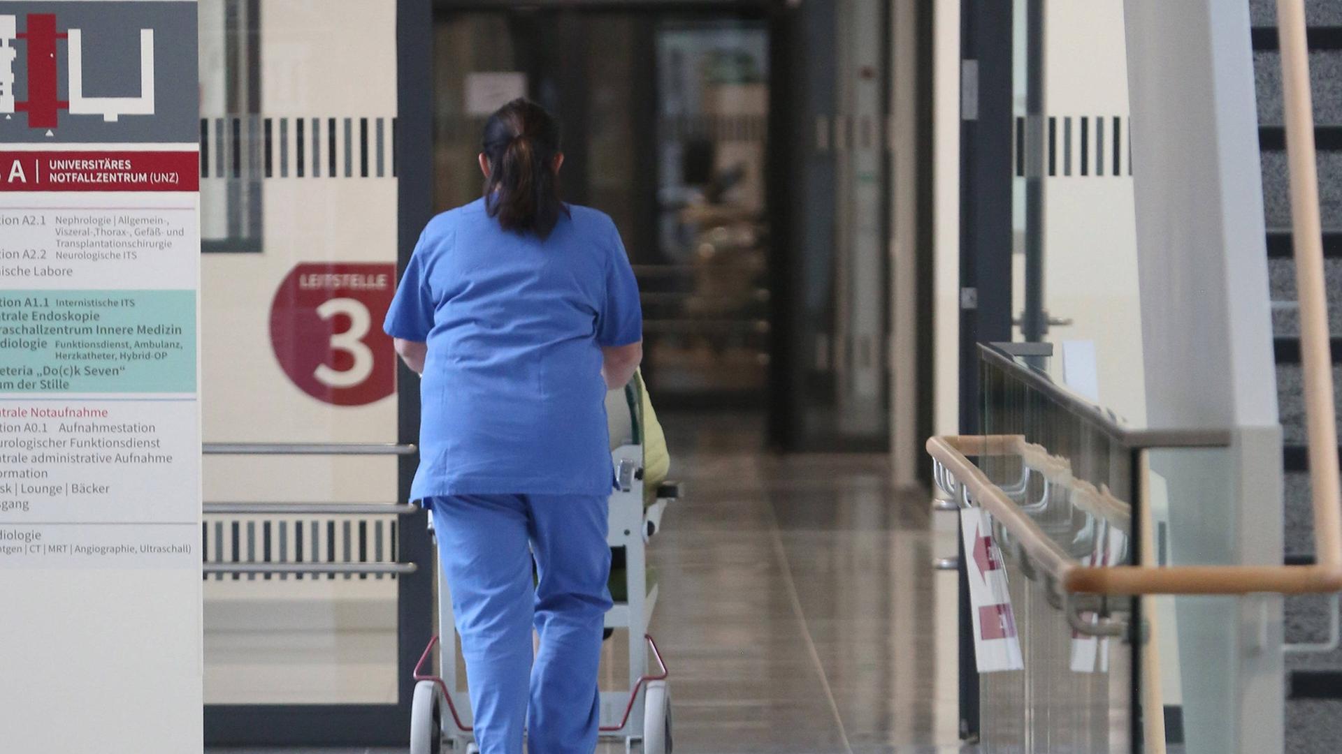 Eine Pflegekraft in blauer Pflegekleidung schiebt eine Person in einem Rollstuhl im Universitären Notfallzentrum in der Hanse- und Universitätsstadt Rostock.