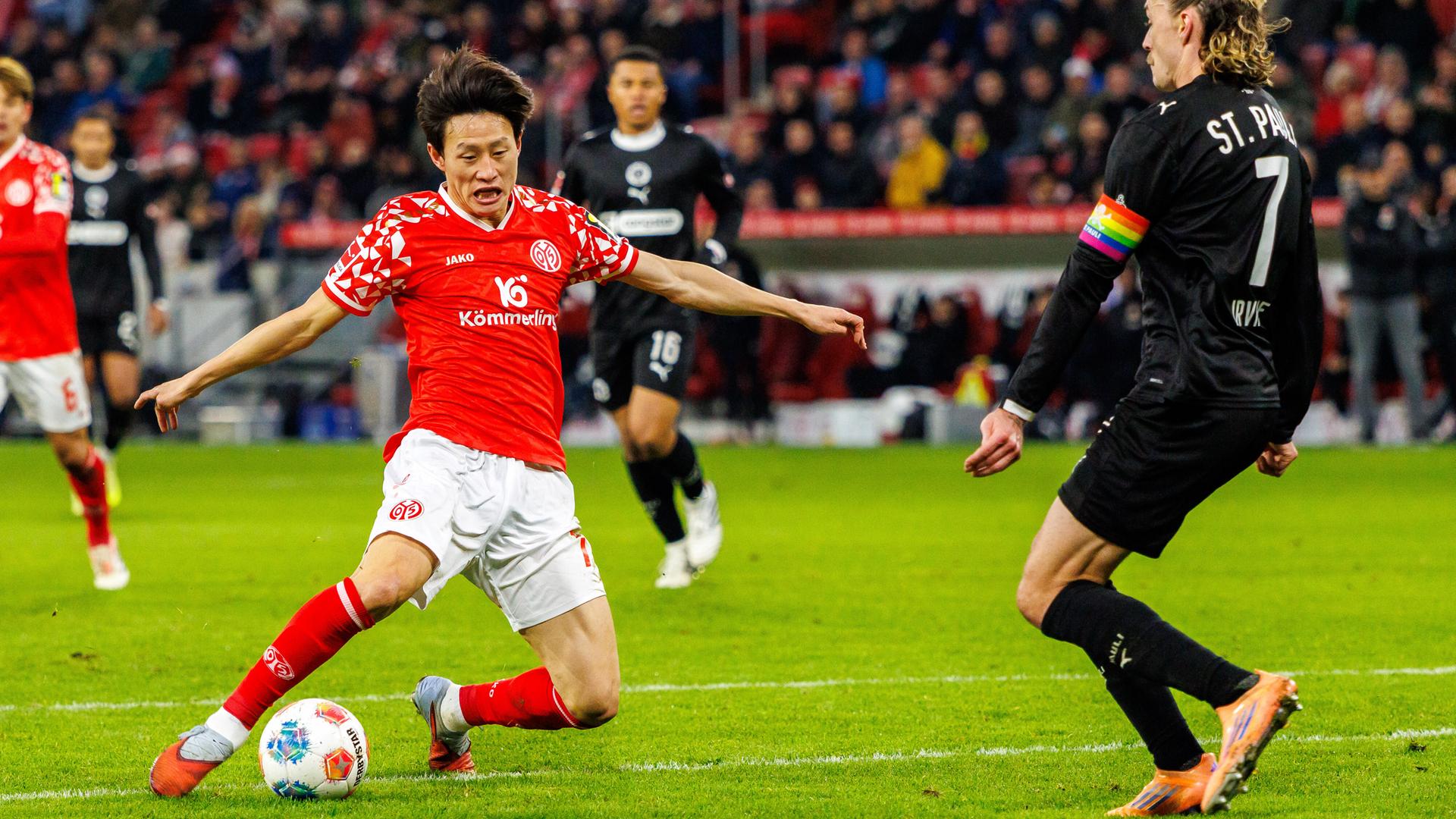 Jae-sung Lee trägt ein rotes Trikot und schießt den Ball auf dem Spielfeld. Jackson Irvine trägt ein schwarzes Trikot und steht ihm gegenüber.