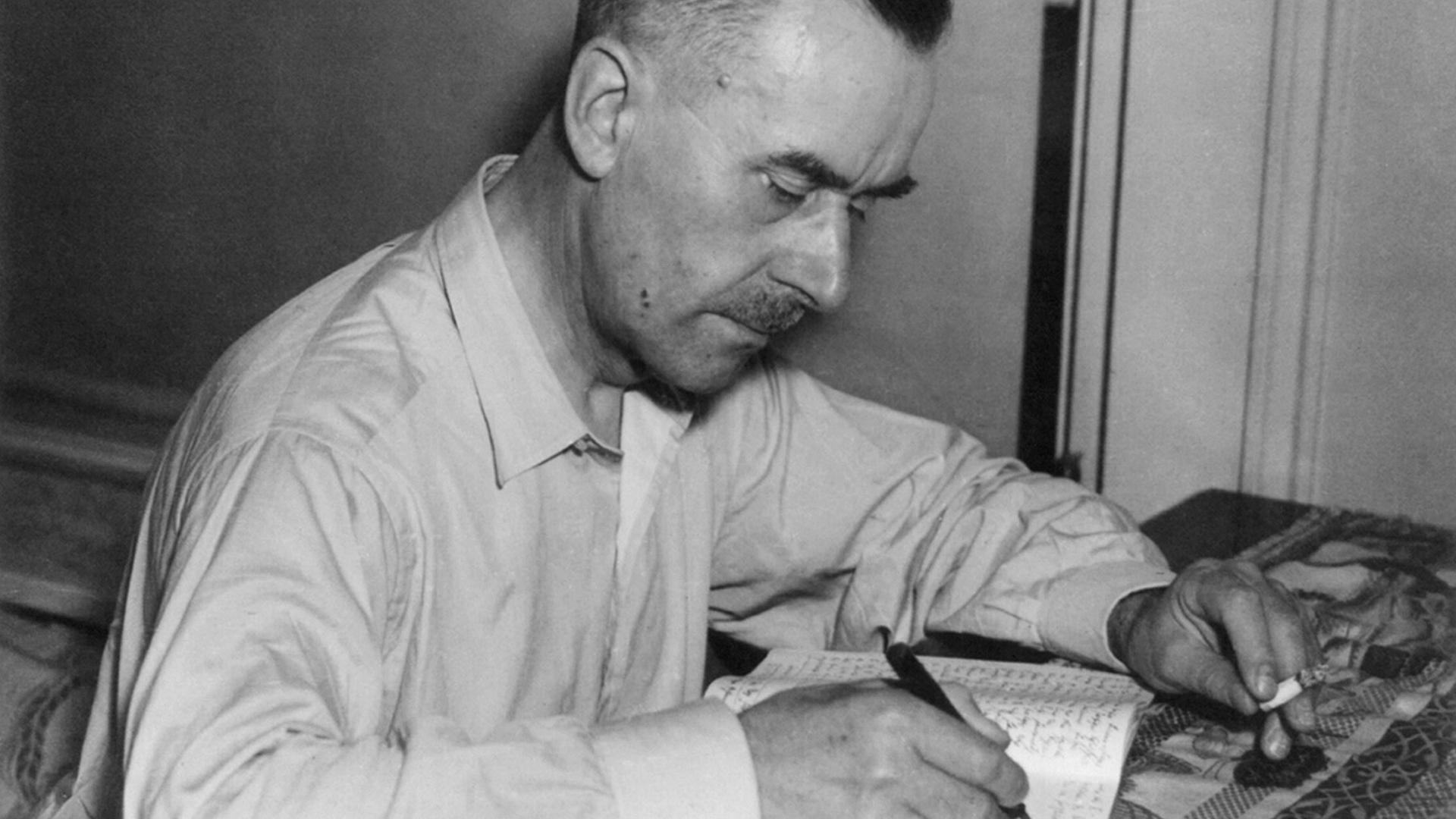 Der Schriftsteller Thomas Mann sitzt an einem Tisch und schreibt in ein Notizbuch. In der anderen Hand hält er eine Zigarette.