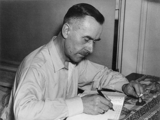 Der Schriftsteller Thomas Mann sitzt an einem Tisch und schreibt in ein Notizbuch. In der anderen Hand hält er eine Zigarette.