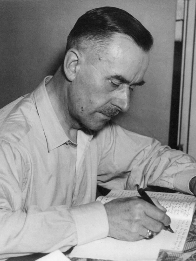 Der Schriftsteller Thomas Mann sitzt an einem Tisch und schreibt in ein Notizbuch. In der anderen Hand hält er eine Zigarette.
