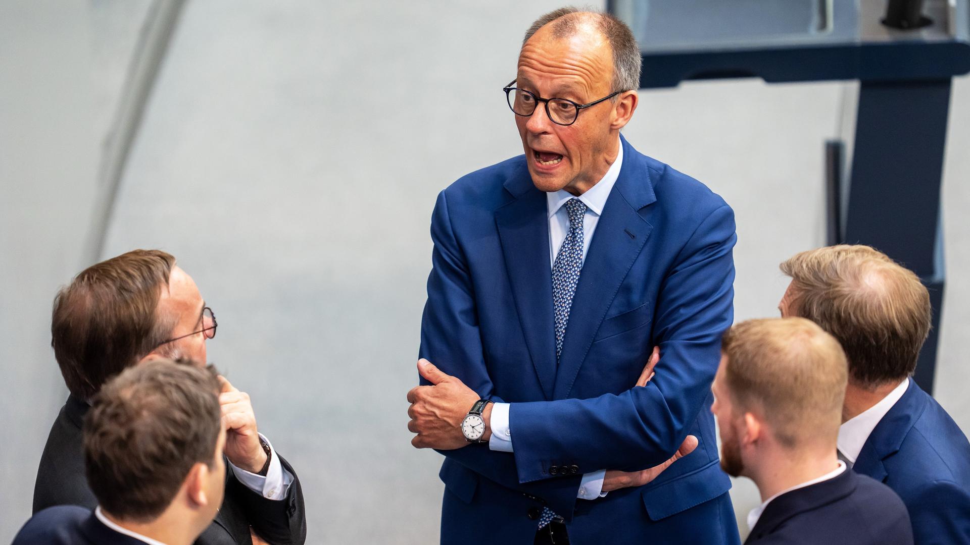 Bundeskanzler Merz spricht im Bundestag mit Ministern von der SPD und der CDU. Sie stehen im Bundestag. Merz hat die Arme verschränkt und den Mund weit aufgerissen. Bundeskanzler Merz spricht im Bundestag mit Ministern von der SPD und der CDU. Sie stehen im Bundestag. Merz hat die Arme verschränkt und den Mund weit aufgerissen.