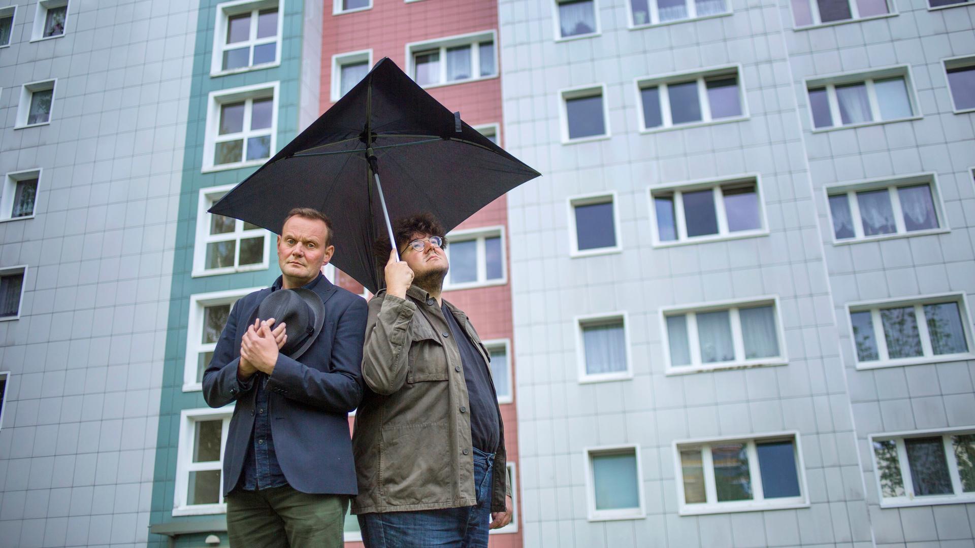 Devid Striesow und Axel Ranisch stehen unter einem schwarzen quadratischen Regenschirm vor der Fassade eines Hochhauses und schauen besorgt