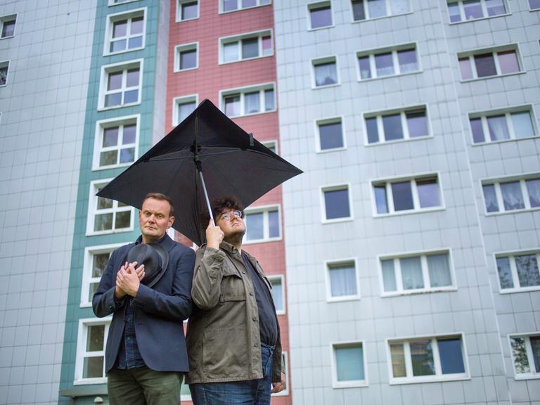 Devid Striesow und Axel Ranisch stehen unter einem schwarzen quadratischen Regenschirm vor der Fassade eines Hochhauses und schauen besorgt