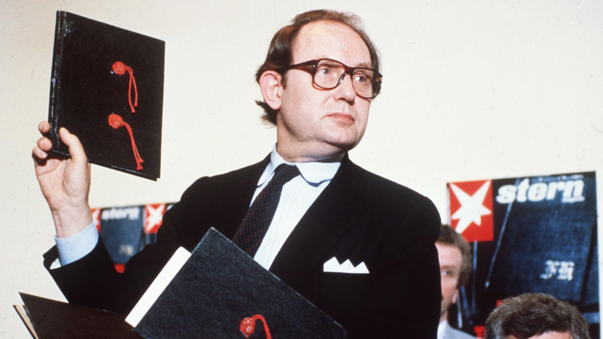 Der Journalist Gerd Heidemann, im schwarzen Anzug mit Krawatte und Einstecktuch, hält eine schwarze Kladde mit zwei roten Siegeln in die Höhe.
