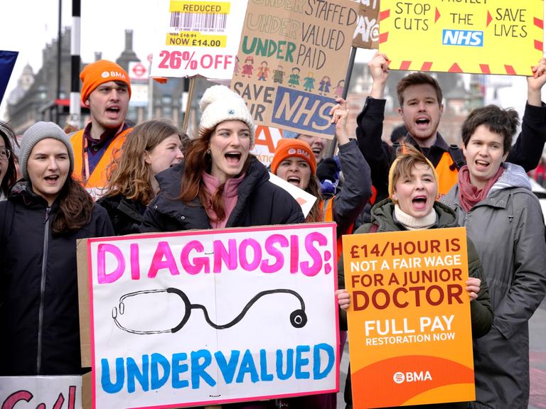 Die Männer und Frauen halten Transparente und Plakate hoch. Auf einem steht "Diagnosis Undervalued".