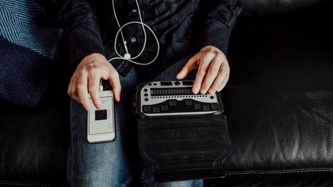 Bild einer Person, die von oben zu sehen ist. Auf einem Knie liegt ein Smartphone, auf dem anderen Knie eine Braille Tastatur