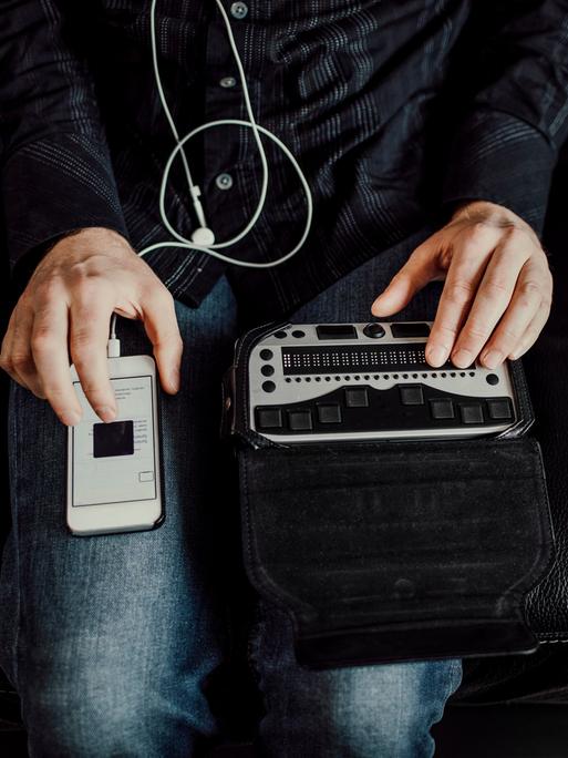 Bild einer Person, die von oben zu sehen ist. Auf einem Knie liegt ein Smartphone, auf dem anderen Knie eine Braille Tastatur