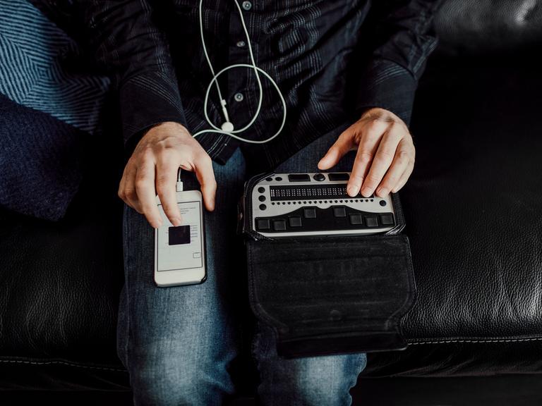 Bild einer Person, die von oben zu sehen ist. Auf einem Knie liegt ein Smartphone, auf dem anderen Knie eine Braille Tastatur