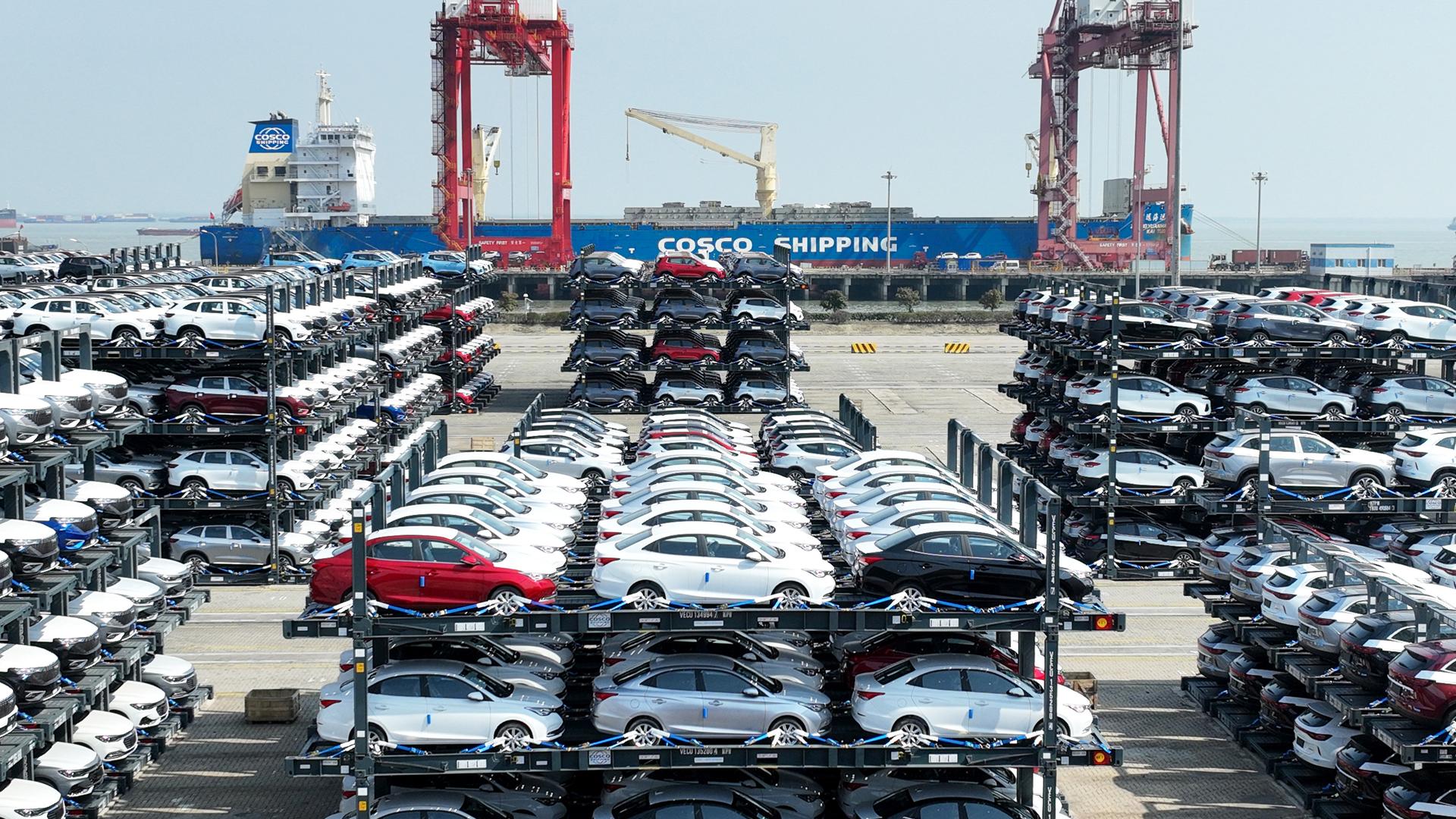 Tausende Autos stehen im Taicang Port International Container Terminal bei Schanghai in spezillen Transportrahmen. Im Hintergrund ein Schiff der Cosco-Reederei und Kräne.