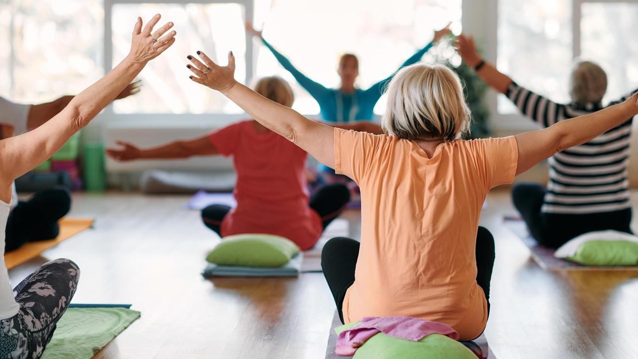 Eine Gruppe älterer Frauen macht Yoga in einem sonnendurchfluteten Raum, alle strecken die Arme in die Luft.