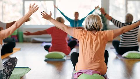 Eine Gruppe älterer Frauen macht Yoga in einem sonnendurchfluteten Raum, alle strecken die Arme in die Luft.
