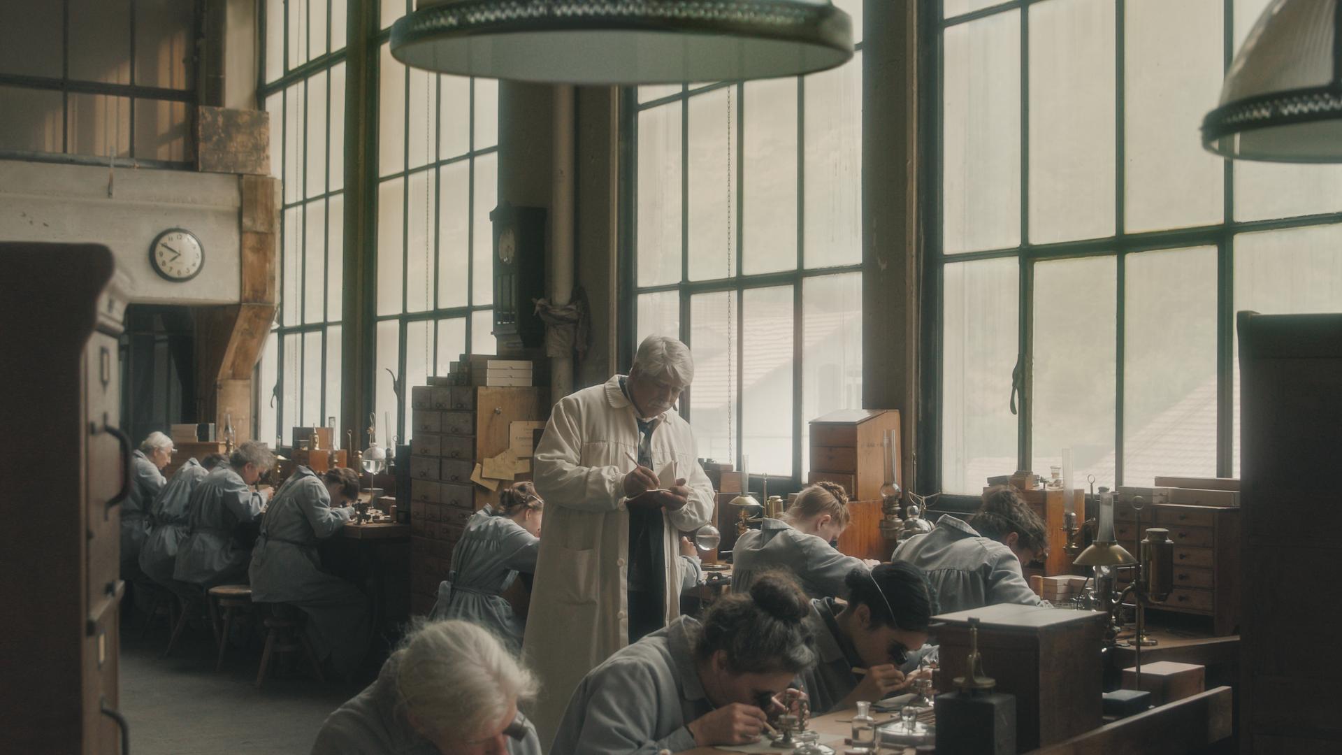 Der Filmausschnitt zeigt Arbeiterinnen in einer Uhrenfabrik, die heruntergebeugt an Tischen arbeiten, hinter ihnen steht ein Mann im weißen Kittel und kontrolliert sie.