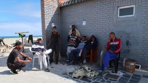 Eine Gruppe junger schwarzer Männer in Trainings- und Freizeitkleidung hat sitzt im Schatten einer Hausecke, im Hintergrund ist ein Sandstrand zu sehen.