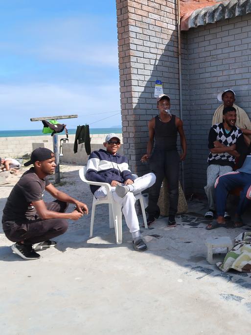 Eine Gruppe junger schwarzer Männer in Trainings- und Freizeitkleidung hat sitzt im Schatten einer Hausecke, im Hintergrund ist ein Sandstrand zu sehen.