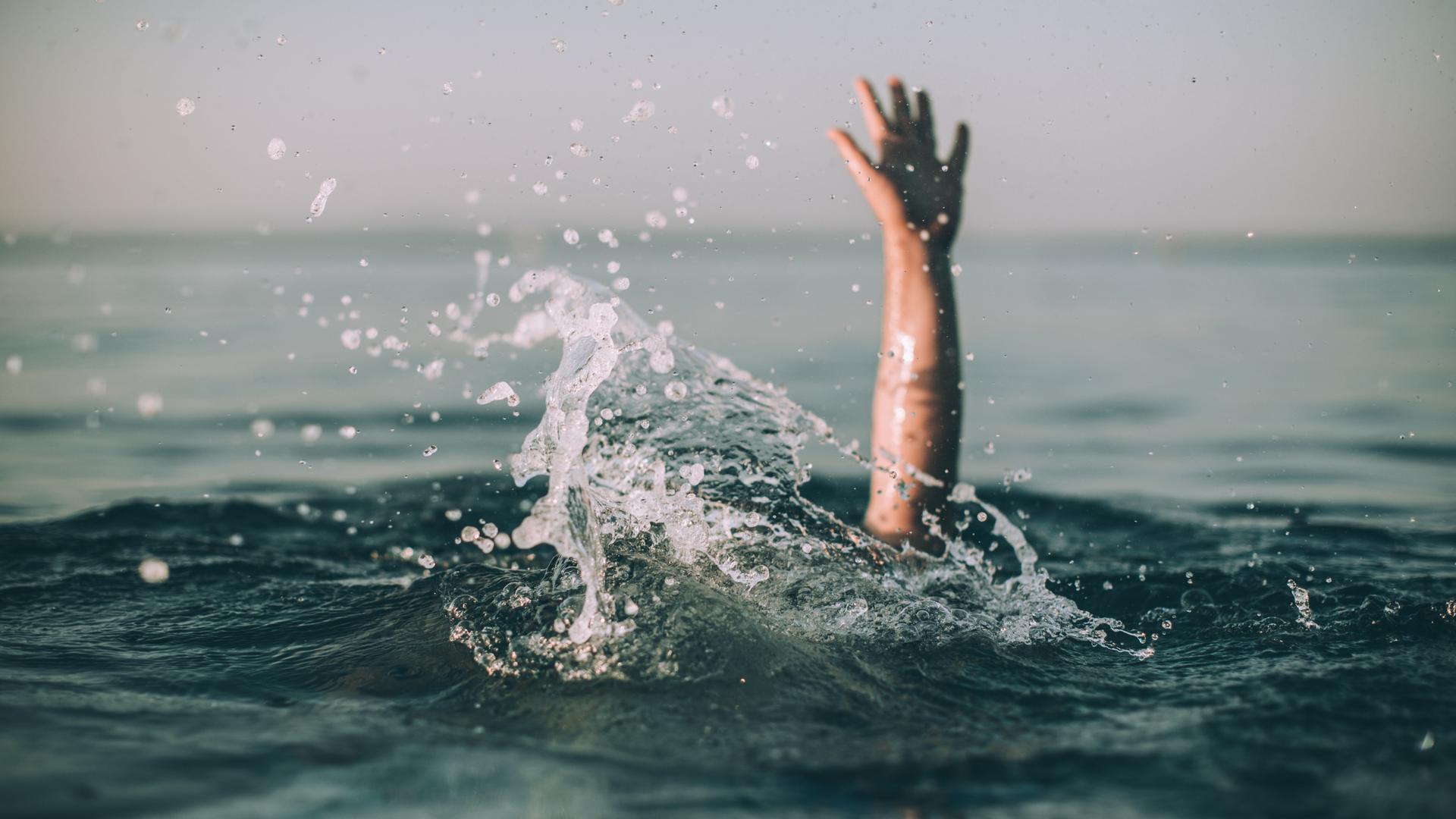 Eine winkende Hand ragt aus einer aufgewühlten Wasseroberfläche hervor. Eine winkende Hand ragt aus einer aufgewühlten Wasseroberfläche hervor.