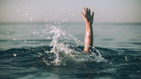 Eine winkende Hand ragt aus einer aufgewühlten Wasseroberfläche hervor.
