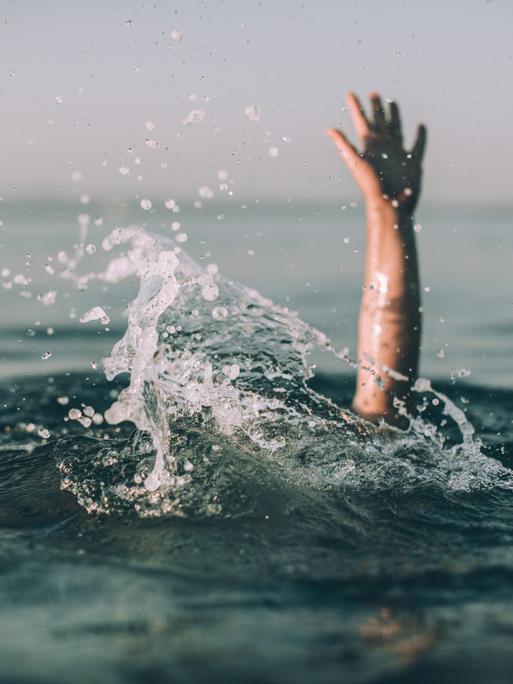 Eine winkende Hand ragt aus einer aufgewühlten Wasseroberfläche hervor.