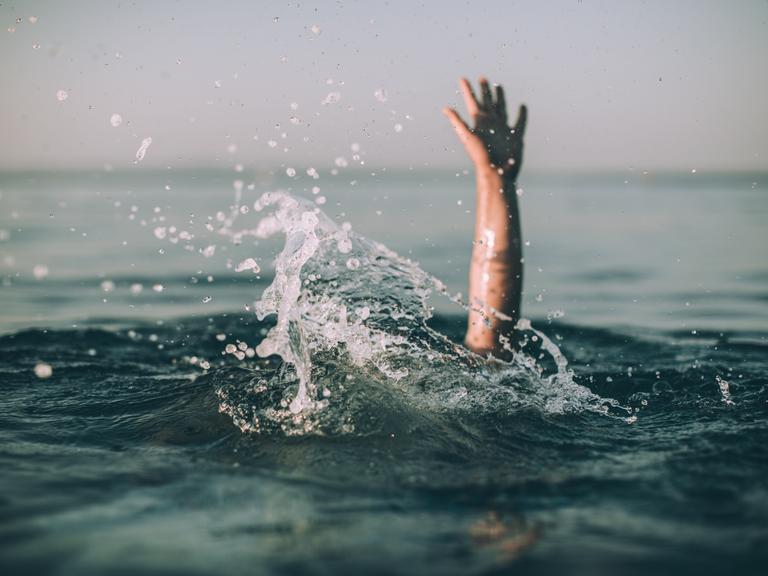 Eine winkende Hand ragt aus einer aufgewühlten Wasseroberfläche hervor.