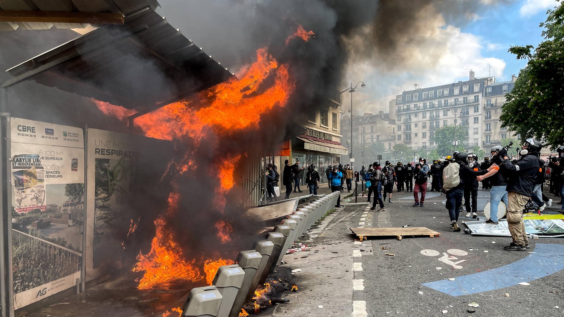 In der Nähe des Place de La Nacion in Paris brennt eine Bushaltestelle. Auf der Straße sind dunkel gekleidete Menschen, teilweise mit Gasmasken, zu sehen.