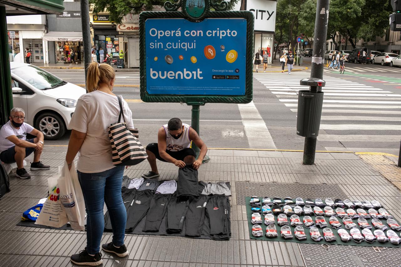 Ein Straßenhändler hockt auf der Straße und verkauft Hosen und Socken. Hinter ihm steht ein Schild mit Bitcoin-Werbung.