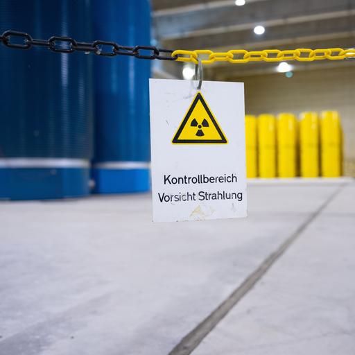 Gelbe und blaue Castor-Behälter stehen penibel gestapelt in einer großen, hellen unterirdischen Halle. Auf einem Schild im Bildvordergrund steht der Hinweis "Kontrollbereich - Vorsicht Strahlung"
