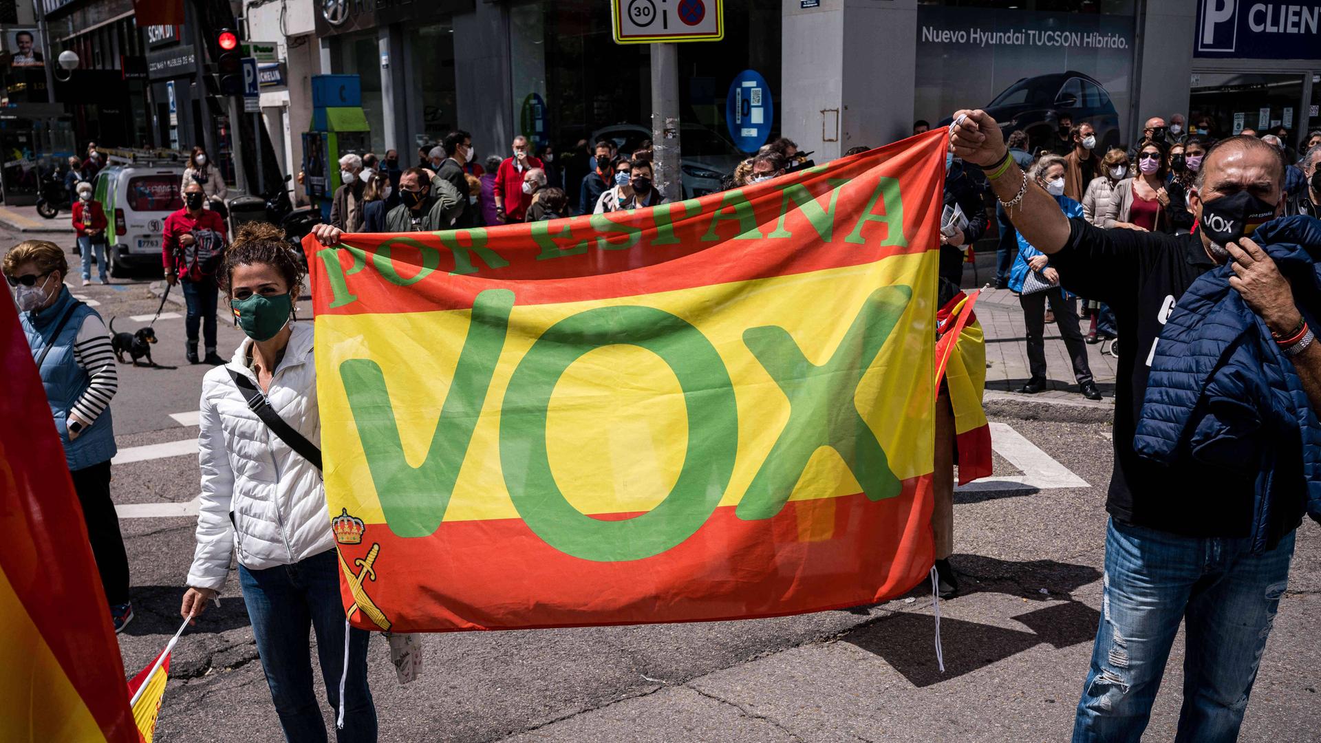 Auf einer Straße halten zwei Demonstranten ein großes buntes Vox-Plakat in die Höhe.
