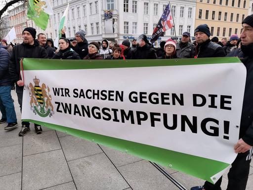 Teilnehmer einer Demonstration halten ein Banner mit der Aufschrift "Wir Sachsen gegen die Zwangsimpfung!", daneben ist das Wappen des Bundeslandes Sachsen abgebildet. 