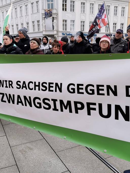 Teilnehmer einer Demonstration halten ein Banner mit der Aufschrift "Wir Sachsen gegen die Zwangsimpfung!", daneben ist das Wappen des Bundeslandes Sachsen abgebildet. 