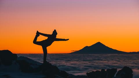 Eine junge Frau macht in Neuseeland Yogaübungen bei Sonnenuntergang über den Wolken, im Hintergrund ist der Mount Taranaki zu sehen.

