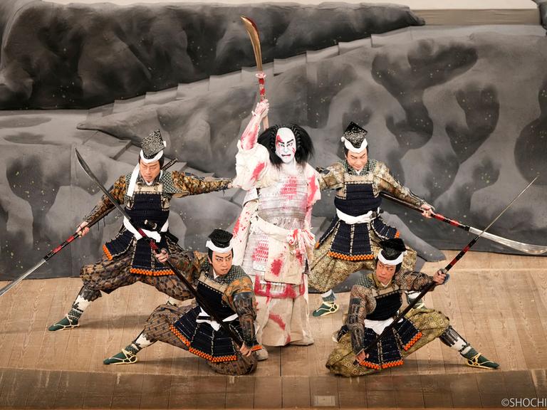 Das Bild zeigt japanische Schauspieler in einer Kabuki-Aufführung. In der Mitte steht ein Krieger mit weißer Maske und weißer, blutverschmierter Kleidung. Er hält ein Schwert in der erhobenen Hand und ist umringt von Kriegern in dunkler Kleidung. 