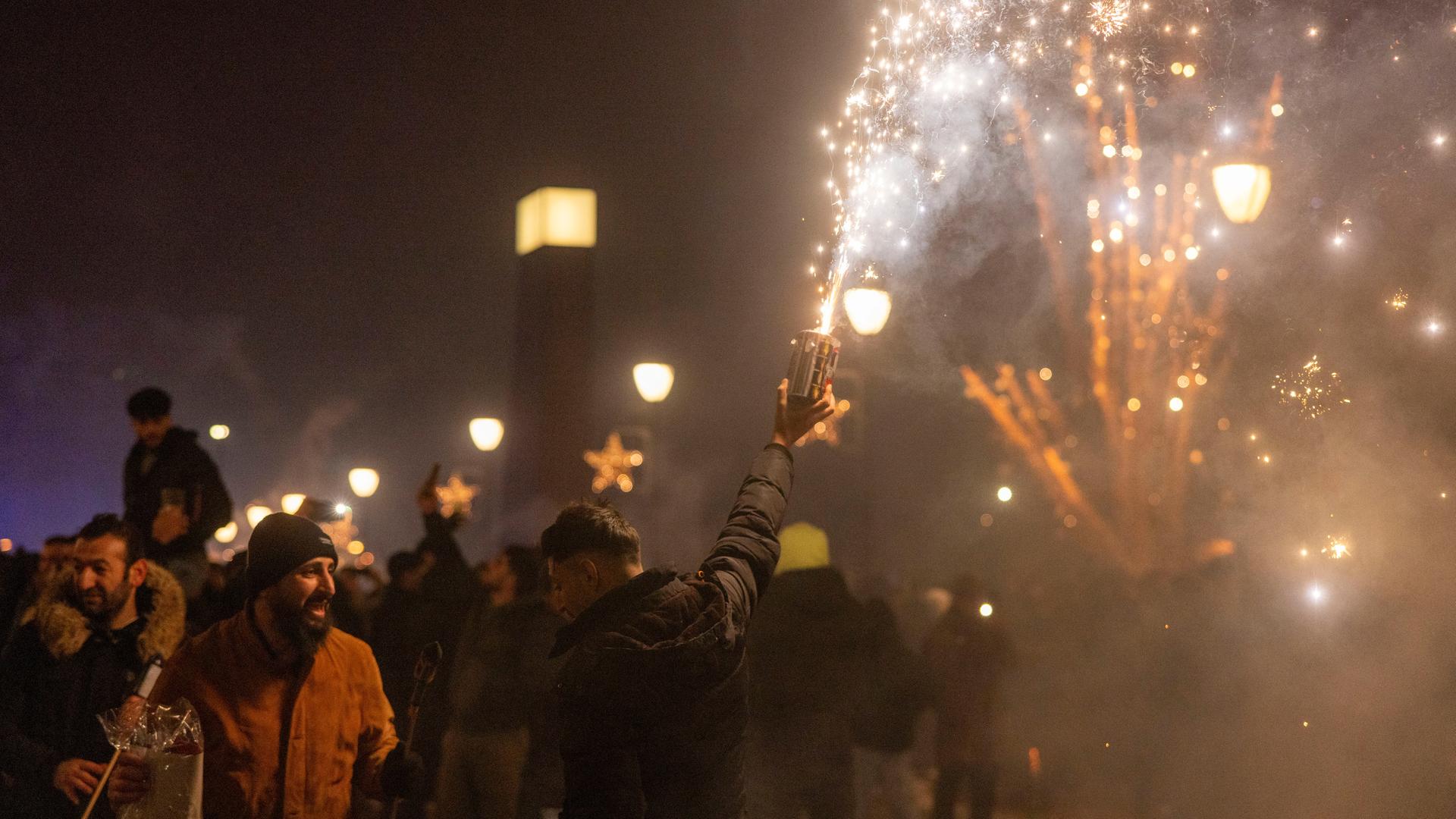 Menschen feiern den Jahreswechsel an Silvester in Frankfurt. Ein Mann hält eine Feuerwerksbatterie in der Hand. Auch im Hintergrund ist Feuerwerk zu sehen.