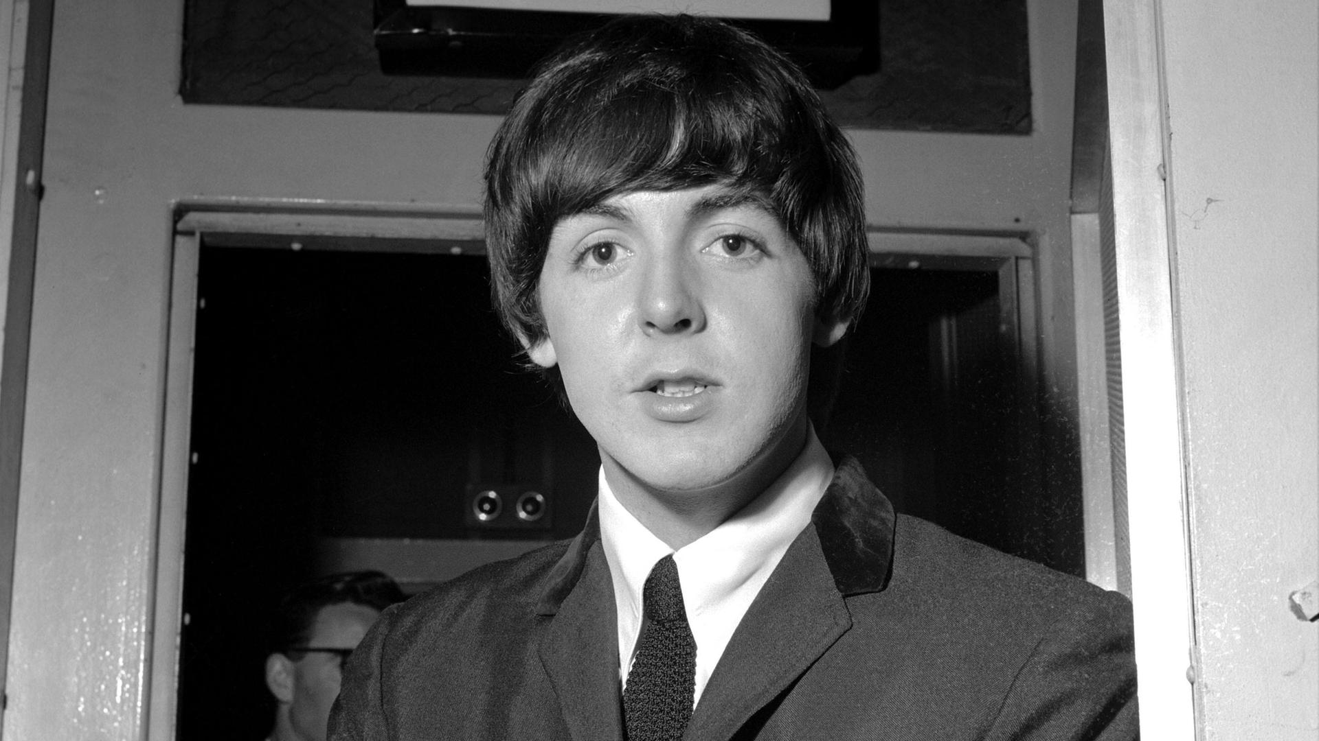 Eine alte Schwarzweißaufnahme von 1964 zeigt den jungen Paul McCartney, damals Sänger und Bassist bei den Beatles. Er trägt die typische Pilzkopffrisur mit Pony und Anzug und Krawatte.