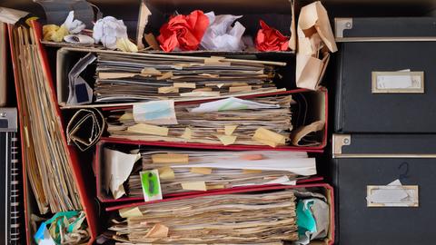Ein Regal voll von unordentlichen Aktenordnern, alten Papieren, zerknüllten Papierkugeln und Kisten.