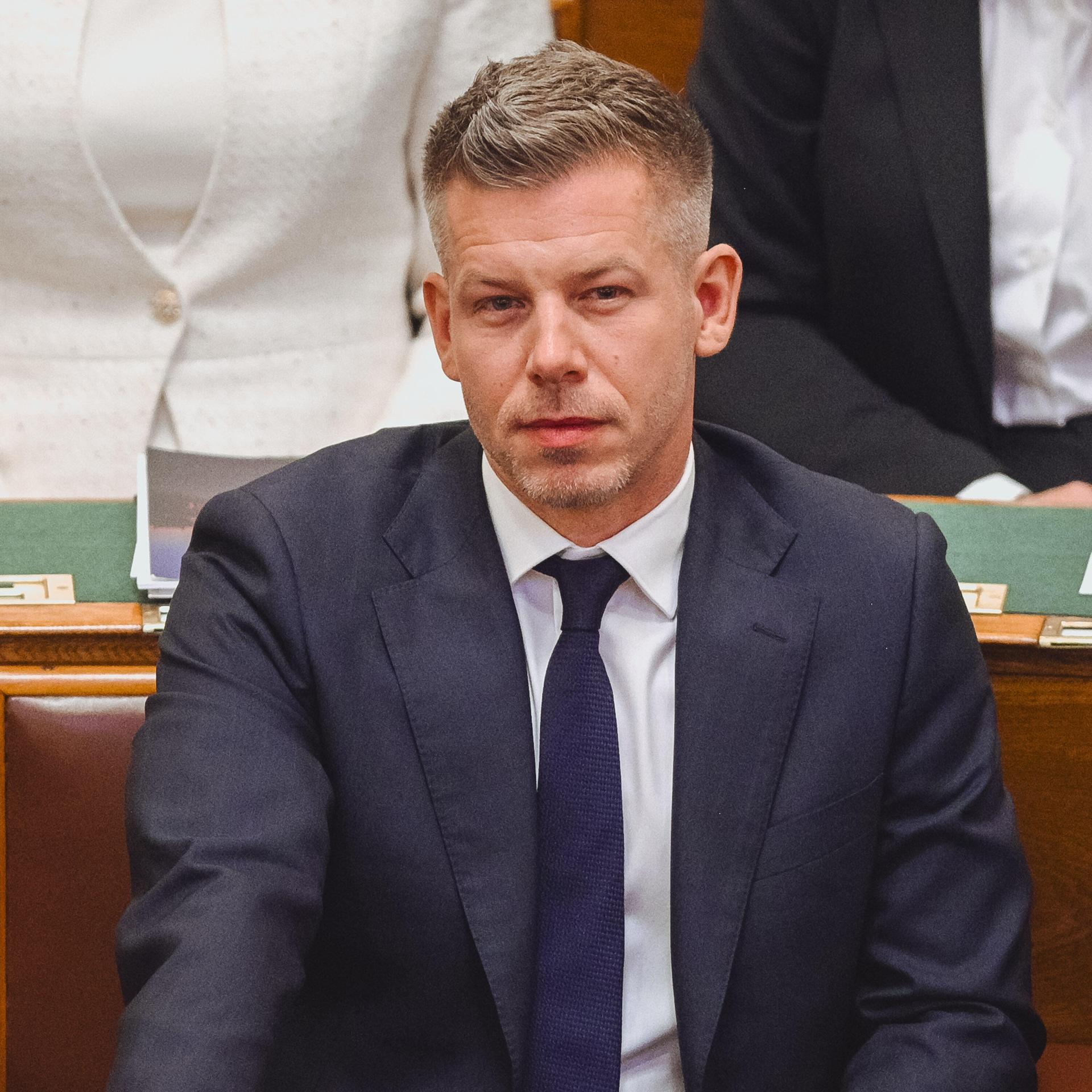 Peter Magyar sitzt auf einer Bank aus Holz. Er trägt einen Anzug und lächelt. Hinter ihm sitzen andere Menschen.