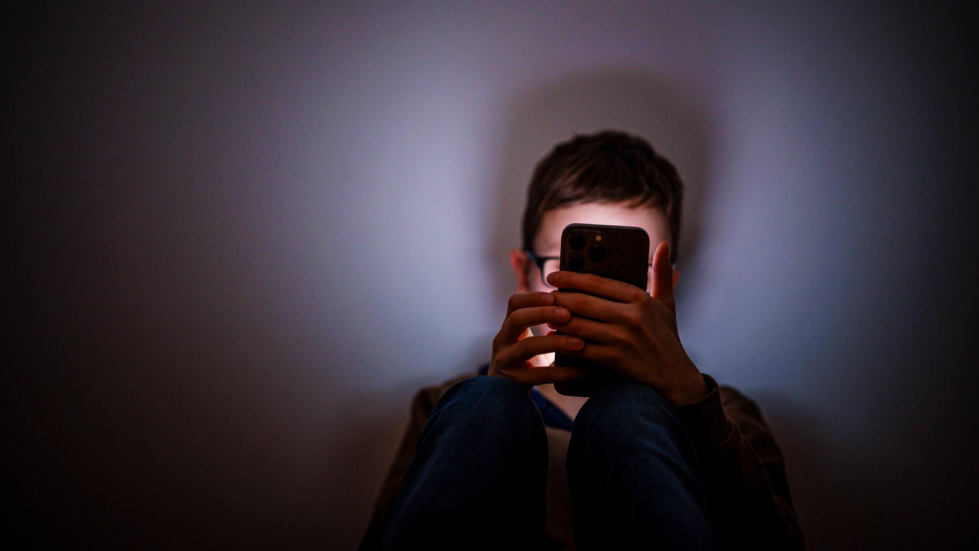 Ein Junge sitzt in einem dunklen Raum und wird von einem Smartphone beleuchtet.