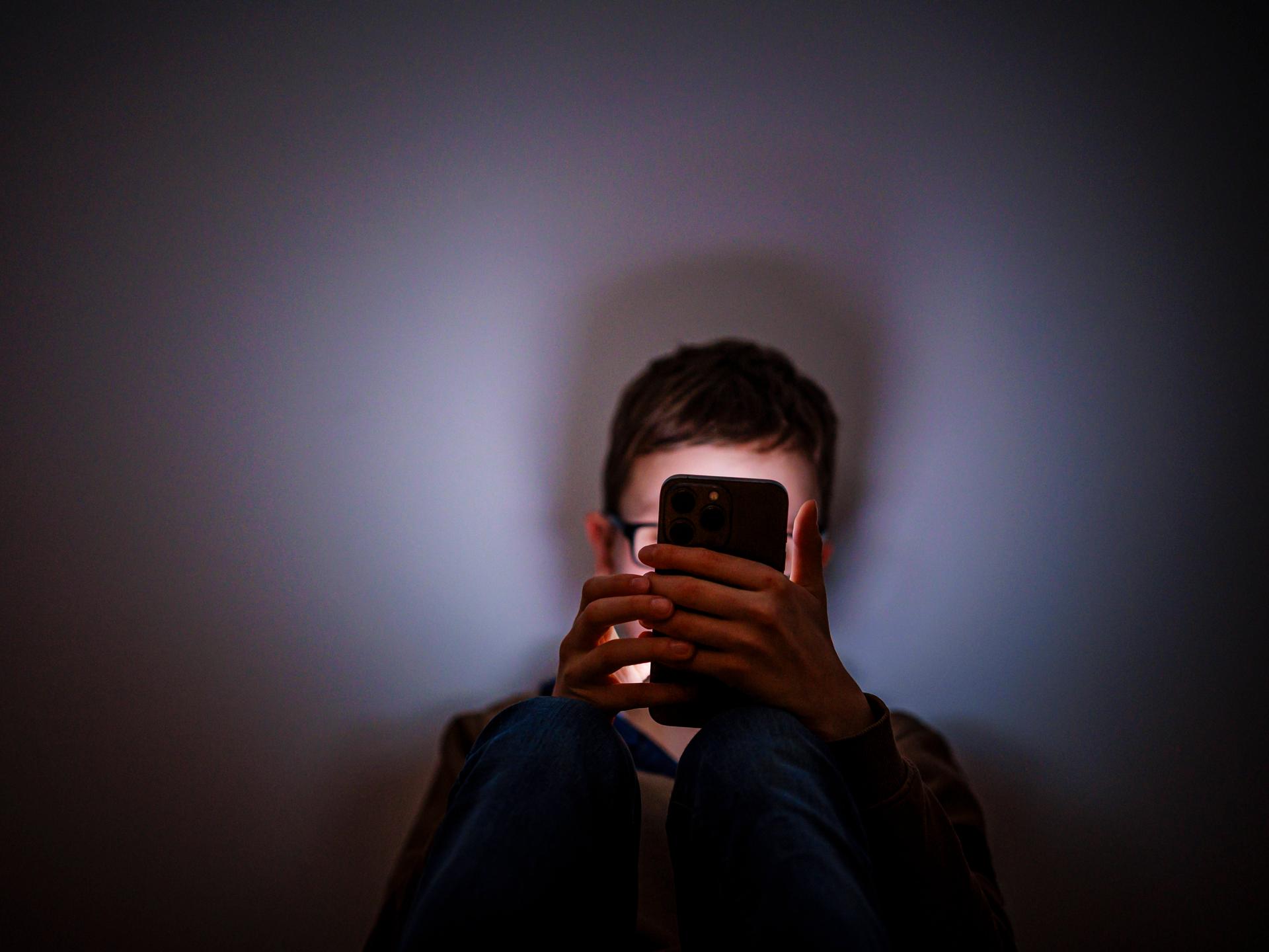 Ein Junge sitzt in einem dunklen Raum und wird von einem Smartphone beleuchtet.