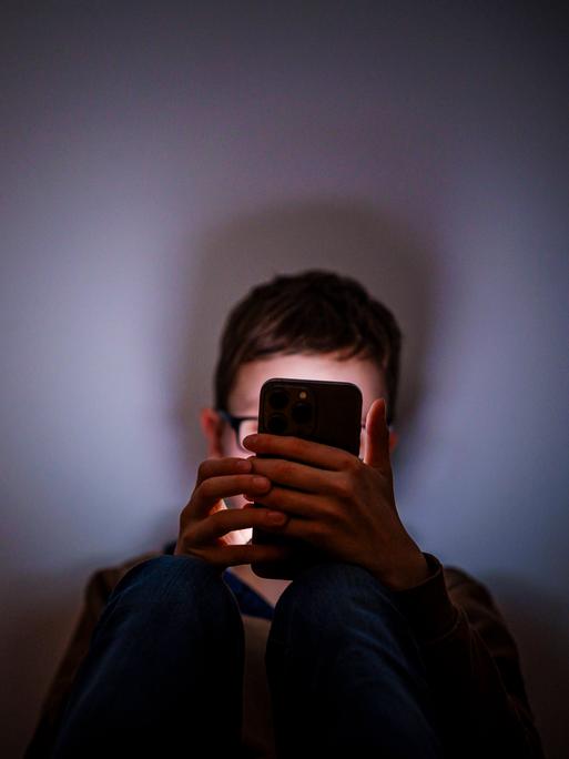 Ein Junge sitzt in einem dunklen Raum und wird von einem Smartphone beleuchtet.